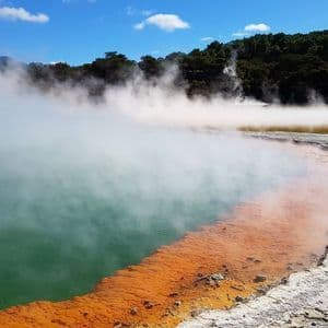 De la vapeur s'élève d'une piscine géothermique aux eaux vertes et oranges vives, bordée par une rive rocheuse avec une forêt en arrière-plan.