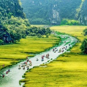 Una vista aerea di molte piccole imbarcazioni che navigano un fiume che serpeggia tra vaste risaie verdi e tra montagne lussureggianti e ripide.