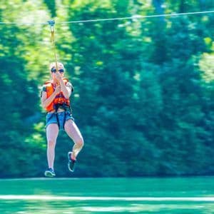 Una donna con giubbotto di salvataggio e occhiali da sole sfreccia in zip-line sopra uno specchio d'acqua, di fronte a una foresta verde.