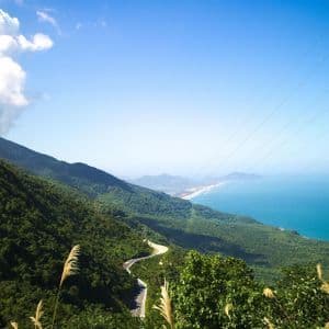 Vista panoramica di una strada tortuosa su una montagna verdeggiante che domina una costa sabbiosa e il mare blu.