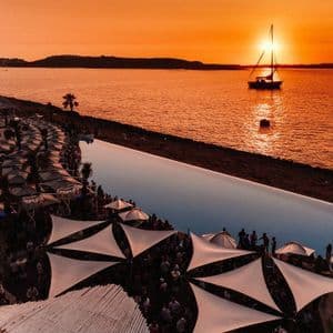 Luftaufnahme einer lebhaften Party an einem Küstenort mit Infinity-Pool, dazu ein Segelboot auf dem Wasser bei orangefarbenem Sonnenuntergang.