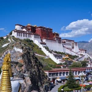 Un grande edificio bianco e rosso su una collina sotto un cielo blu, con uno stupa dorato in primo piano e montagne sullo sfondo.