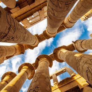 Una vista en contrapicado de grandes columnas de piedra con jeroglíficos antiguos que se elevan hacia un cielo azul y nublado.