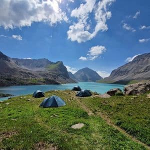 Vier blaue Zelte auf einem grasbewachsenen Hügel mit Blick auf einen türkisfarbenen Bergsee und Berge.