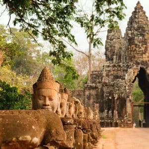 A row of weathered stone statues with carved faces leads to an ancient, ornate stone gate under lush green trees.