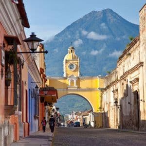Un arco giallo con una torre dell'orologio si erge su una strada di ciottoli, con un grande vulcano che si staglia sullo sfondo.