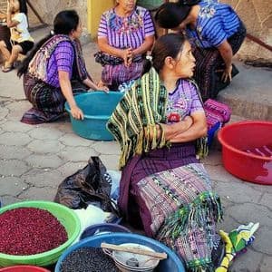 Un gruppo di donne in abiti tradizionali siede su una strada lastricata vendendo fagioli colorati da grandi bacinelle.