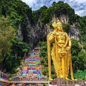 Una gigantesca estatua dorada se erige junto a una larga escalera de colores arcoíris que asciende por un exuberante acantilado verde, mientras la gente sube.