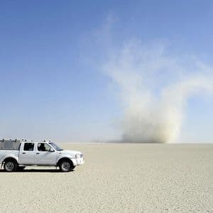 Un pick-up bianco è parcheggiato su una vasta, piatta pianura desertica mentre un grande diavolo di sabbia vortica in lontananza sotto un cielo azzurro e limpido.