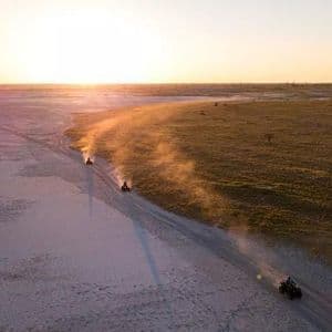 Una vista aerea di un viaggio di gruppo WeRoad in quad su una pista sterrata attraverso un vasto paesaggio al tramonto, sollevando polvere.