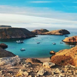 Una vista aerea di diverse barche ancorate in una baia turchese, circondata da aride colline rocciose sotto un cielo azzurro.