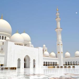 Una grande moschea bianca con cupole multiple e un minareto, vista da un cortile di marmo riflettente sotto un cielo azzurro limpido.