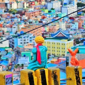 Una statuetta del Piccolo Principe e una volpe siedono su dissuasori gialli, affacciandosi su un paesaggio urbano denso e colorato.