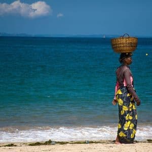 Eine Frau geht an einem Sandstrand am blauen Ozean entlang und balanciert einen großen Weidenkorb auf ihrem Kopf unter klarem Himmel.