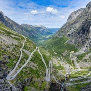 Una vista aerea di una strada tortuosa con tornanti che scende attraverso una ripida e verde valle di montagna sotto un cielo parzialmente nuvoloso.
