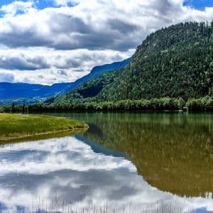 La surface calme d'un lac reflète des montagnes vertes et boisées et un ciel partiellement nuageux.