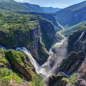 Ein großer Wasserfall stürzt in eine tiefe Schlucht, während ein Fluss durch ein üppiges, grünes Gebirgstal fließt.