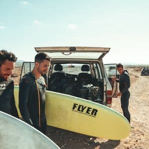 Un viaje en grupo de WeRoad con tres hombres en trajes de neopreno sosteniendo tablas de surf junto a un SUV con el maletero abierto en un paisaje árido.
