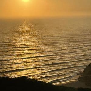 El sol se pone sobre un océano brumoso, proyectando un reflejo dorado en el agua mientras las olas ruedan hacia una orilla silueteada.