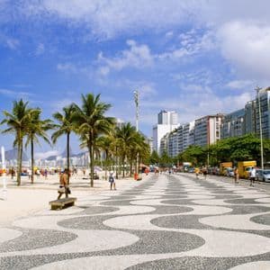 Un paseo adoquinado bordea una playa de arena con palmeras, gente y edificios de la ciudad bajo un cielo azul.