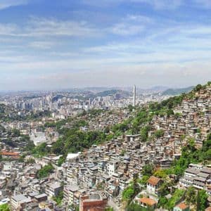 Una vista panoramica di una favela densamente popolata su una verde collina, con lo skyline della città in lontananza.