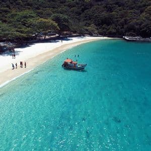 Una vista aerea di una barca su acqua turchese vicino a una cala con spiaggia di sabbia bianca, alberi lussureggianti e persone che nuotano.