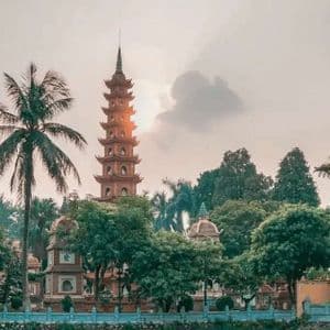 Une tour-pagode orange à plusieurs niveaux est entourée d'arbres verdoyants, y compris des palmiers, sous un ciel voilé au coucher du soleil.