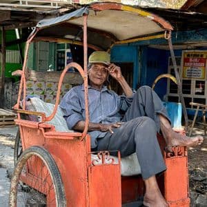 A smiling elderly man with a hat sits barefoot in a traditional orange three-wheeled rickshaw on a sunny street.