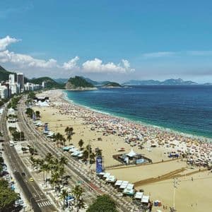 Una vista aérea de una playa concurrida con sombrillas de colores junto a un horizonte urbano y el océano azul bajo un cielo parcialmente nublado.