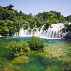 Mehrere breite Wasserfälle stürzen in einen klaren, türkisen Fluss, umgeben von einem dichten grünen Wald unter klarem Himmel.