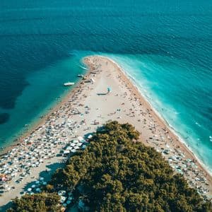 Luftbild einer dreieckigen, mit Menschen und Sonnenschirmen überfüllten Strandzunge, die ins türkisfarbene Wasser ragt und von einem Wald gesäumt wird.