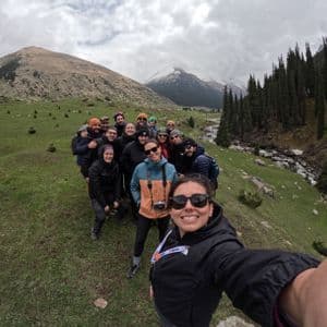 A WeRoad group trip poses for a selfie on a grassy mountainside, with a river and snow-capped peaks in the background.