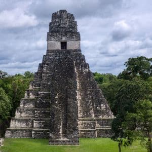 Una gran pirámide escalonada de piedra se alza desde un claro verde en una densa jungla bajo un cielo nublado.