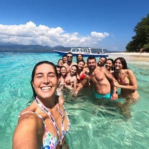 A woman takes a selfie of her WeRoad group trip as they smile together in the clear turquoise water of a tropical beach.