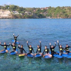 A WeRoad group trip in wetsuits posing on blue surfboards in the clear sea, with a green coastline in the background.