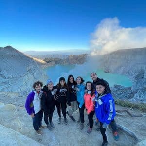 A WeRoad group trip poses for a photo on the edge of a volcanic crater, with a bright turquoise lake in the background.