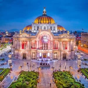 Edificio con cúpula ornamentada iluminado al atardecer, visto desde arriba, con jardines cuidados y una plaza pública en primer plano.