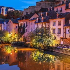 Case a graticcio illuminate e alberi che costeggiano un fiume calmo al crepuscolo, con i loro riflessi visibili nell'acqua.