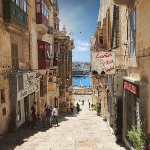 Una vista lungo una ripida e stretta strada di pietra, fiancheggiata da vecchi edifici con balconi, che conduce a un porto azzurro e soleggiato.
