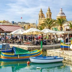 Bateaux colorés amarrés dans un port avec des gens dînant dans des cafés au bord de l'eau sous un ciel ensoleillé.