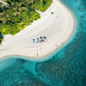 Vue aérienne d'une plage tropicale de sable blanc avec des palmiers, des parasols et des personnes profitant de l'eau turquoise claire.