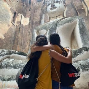 Due donne di un viaggio di gruppo WeRoad si abbracciano mentre guardano una gigantesca statua del Buddha seduto all'interno di un tempio di pietra.