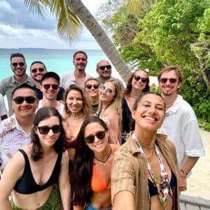 Un groupe WeRoad sourit pour un selfie sur une plage tropicale avec de l'eau turquoise et des palmiers.