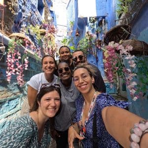 Un voyage de groupe WeRoad de six personnes souriantes prenant un selfie dans une ruelle étroite avec des murs bleu vif et des fleurs suspendues.