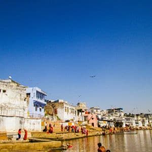 Personas con atuendos coloridos se congregan en ghats de piedra a lo largo de una ribera, con un denso paisaje urbano bajo un cielo azul vibrante.