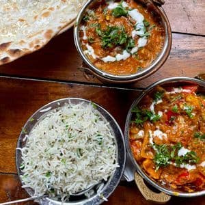 Una vista dall'alto di due ciotole di curry indiano, una ciotola di riso speziato e un pezzo di pane naan su un tavolo di legno.