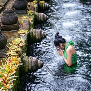 Una donna vestita di verde esegue un rito di purificazione, inchinandosi sotto l'acqua che scorre da un beccuccio di pietra in una vasca sacra.