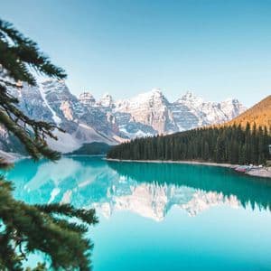 Schneebedeckte Berge und ein Kiefernwald spiegeln sich im türkisfarbenen Wasser eines stillen Sees unter klarem Himmel.