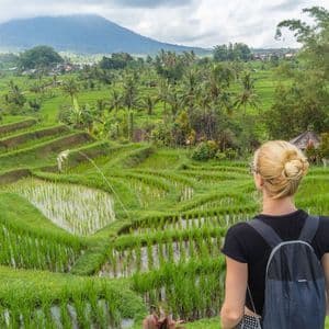 A woman with a backpack looks out over green, terraced rice paddies, with a large mountain in the background under a cloudy sky.