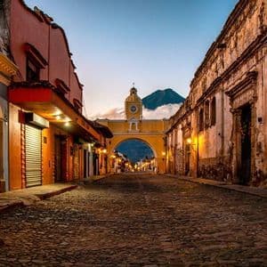 Una strada acciottolata al tramonto conduce a un arco giallo con una torre dell'orologio, con un vulcano visibile sullo sfondo.